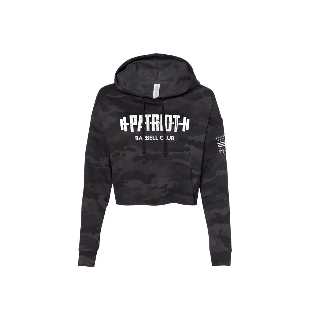 A cropped camo hooded sweatshirt featuring the words Patriot barbell club across the chest. The word PATROT is part of a barbell design. The barbell goes right through the letters, so that the weights are on either side. The sleeve has a distressed flag and Rebecca's signature underneath.