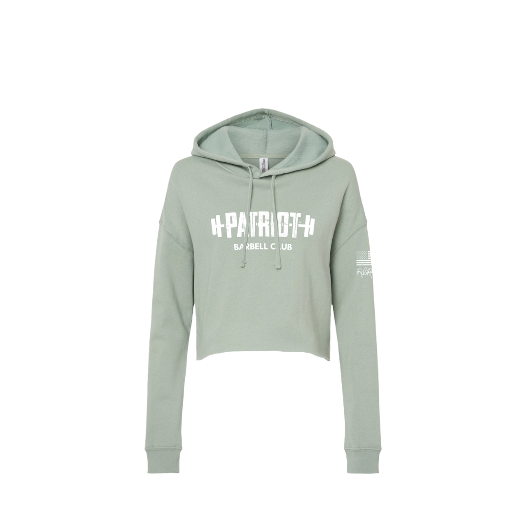 A cropped sage hooded sweatshirt featuring the words Patriot barbell club across the chest. The word PATROT is part of a barbell design. The barbell goes right through the letters, so that the weights are on either side. The sleeve has a distressed flag and Rebecca's signature underneath.
