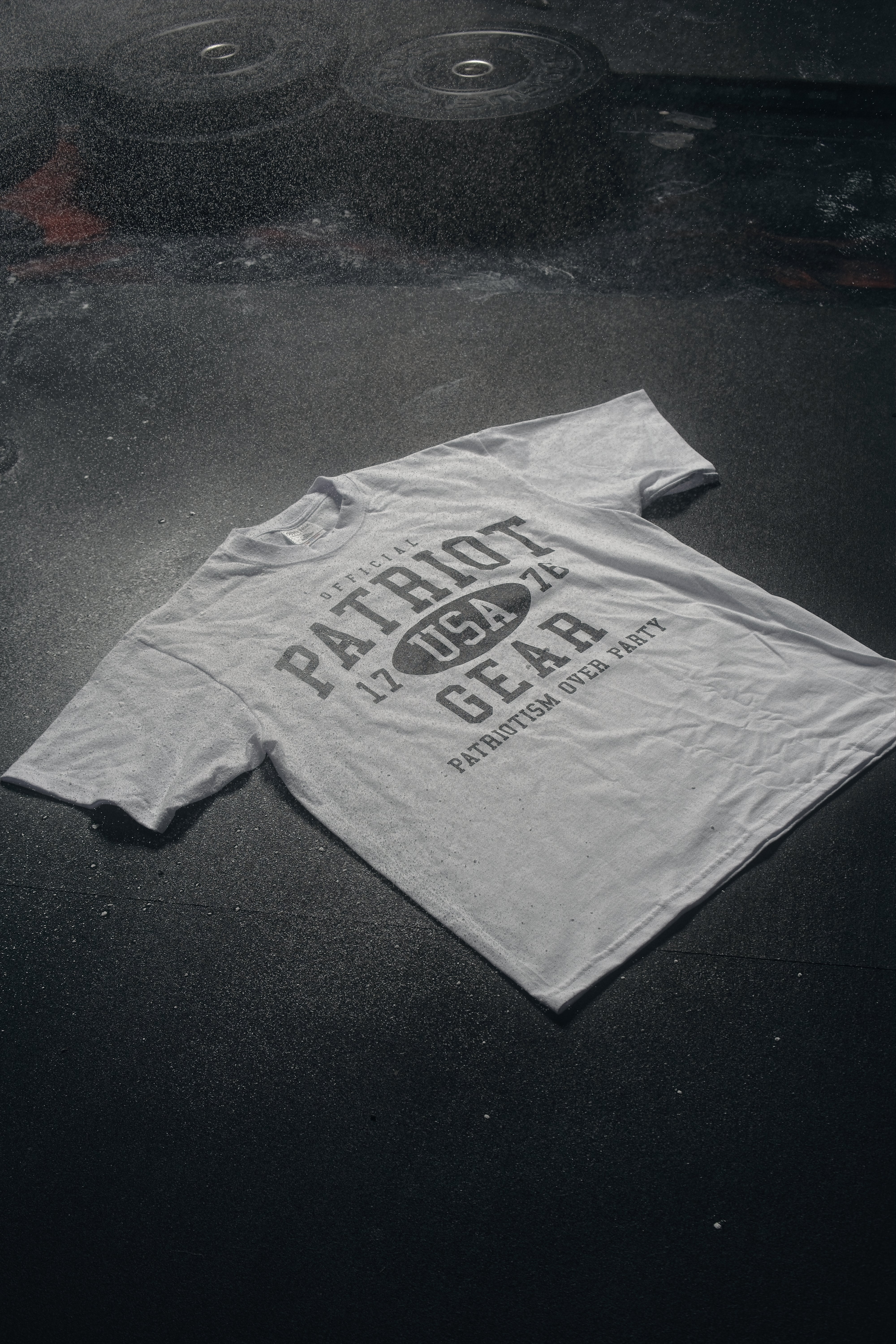 White oversized T-shirt from Official Patriot Gear, on a gym floor. featuring a bold white collegiate-style graphic on the front. The design includes "OFFICIAL PATRIOT GEAR" in large black letters, with "USA" inside an oval between the numbers "17" and "76." Below, the slogan "PATRIOTISM OVER PARTY" is displayed in capital letters. The shirt has a classic crew neck and short sleeves, with a relaxed, athletic fit.
