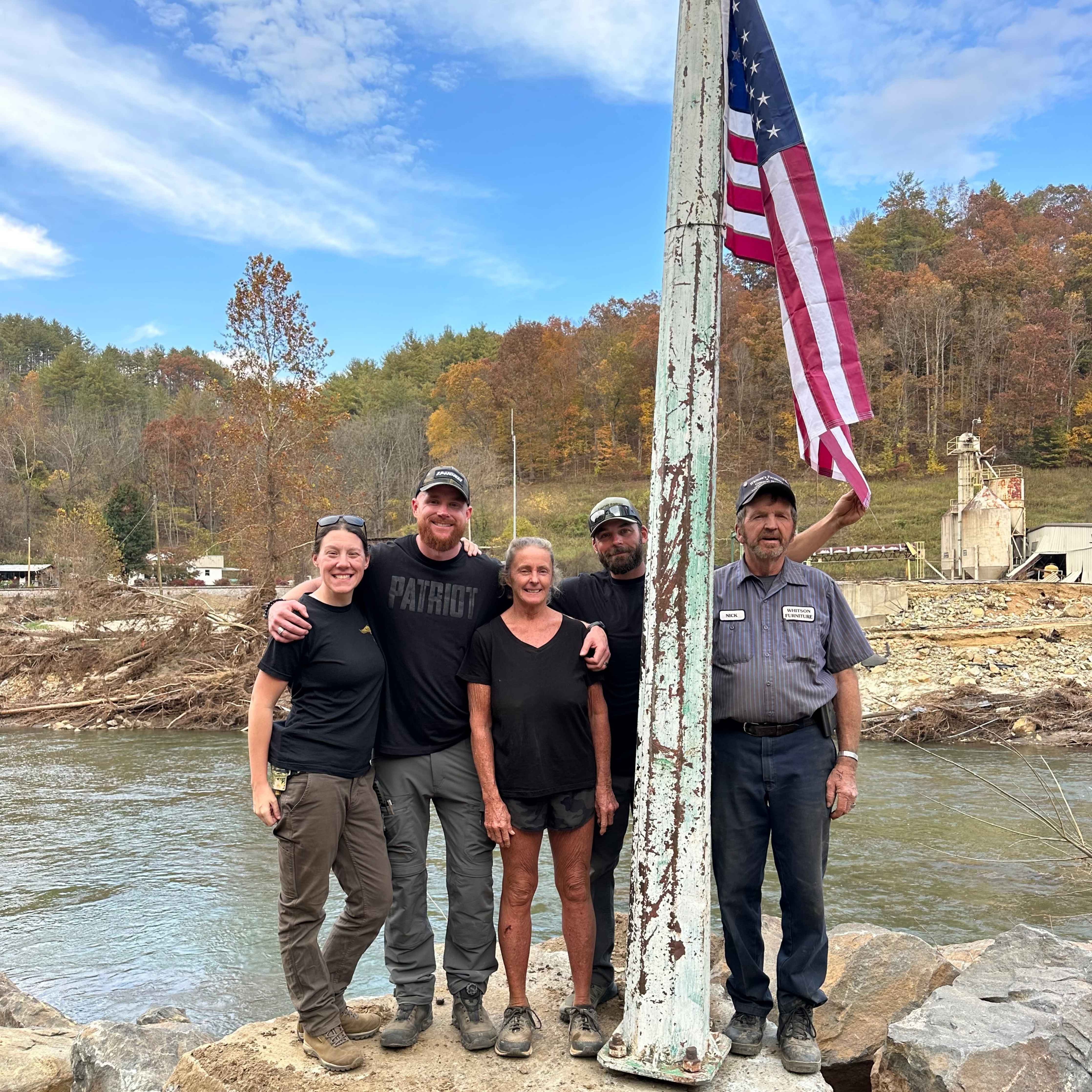 Five people, including OPG team members in branded apparel and local residents, stand together smiling by an American flagpole in a rural area impacted by flooding. 