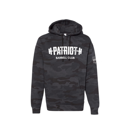 A camo hooded sweatshirt featuring the words Patriot barbell club across the chest. The word PATROT is part of a barbell design. The barbell goes right through the letters, so that the weights are on either side. The sleeve has a distressed flag and Rebecca's signature underneath. 