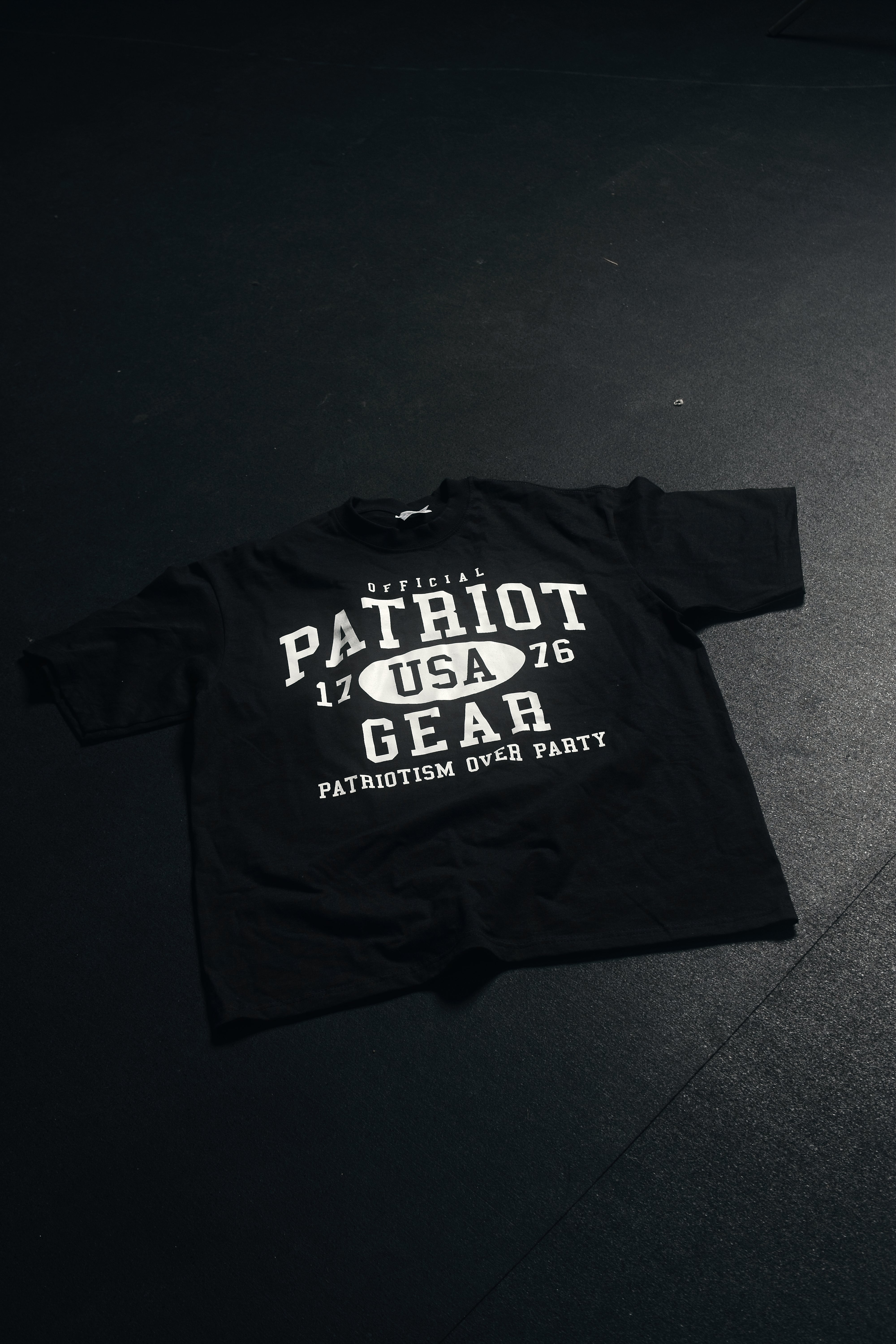 Black oversized T-shirt from Official Patriot Gear, on a gym floor. featuring a bold white collegiate-style graphic on the front. The design includes "OFFICIAL PATRIOT GEAR" in large white letters, with "USA" inside an oval between the numbers "17" and "76." Below, the slogan "PATRIOTISM OVER PARTY" is displayed in capital letters. The shirt has a classic crew neck and short sleeves, with a relaxed, athletic fit. 