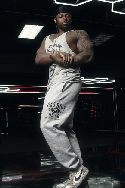 Man flexing, while wearing grey sweatpants with black patriot  gear logo with USA 1776 in the middle and patriotism over party. Decoration is on the side of the leg, under the pocket. 